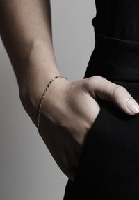 Zlatý řetízkový náramek na zápěstí, skládající se z malých propojených oválních tvarů, ležící na černé látce. Hladká textura pleti je viditelná.