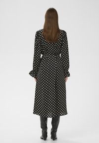 Schwarzes Kleid mit weißen Punkten, langen Ärmeln, tailliert und mit ausgestellten Manschetten; aus leichtem Stoff; kombiniert mit knielangen schwarzen Stiefeln.