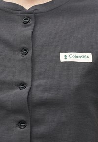 Σκούρο γκρι πουκάμισο με κουμπιά και λογότυπο Columbia Sportswear Company στο στήθος, που δείχνει την υφή του υφάσματος και τέσσερα μαύρα κουμπιά.
