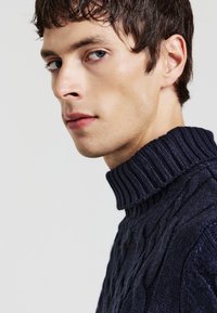 Maglione a collo alto in tessuto a maglia cable color blu navy, con un colletto a coste e un motivo testurizzato. Realizzato in un filato morbido e spesso.