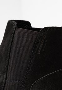 Czarne zamszowe botki za kostkę z elastycznymi panelami bocznymi, tonowanym przeszyciem i subtelnym logo na pięcie. Gładka faktura z eleganckim fasonem.