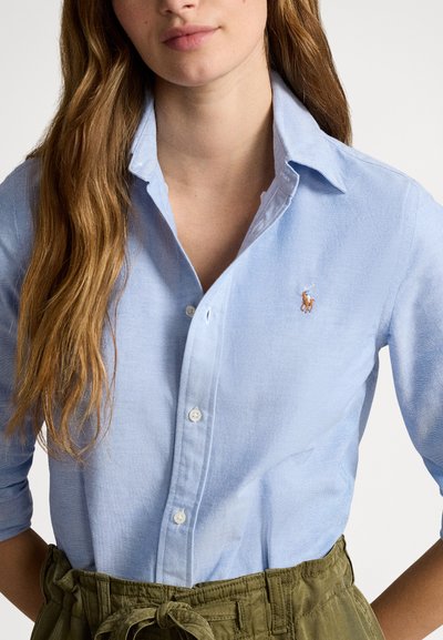 Γυναίκα που φοράει ανοιχτό μπλε πουκάμισο με κουμπιά και μανίκια γυρισμένα, και χακί παντελόνι ψηλής μέσης, δεμένο στη μέση.