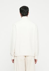 Suéter blanco de punto con cuello alto, dobladillo y puños acanalados. Presenta un ajuste relajado y detalles de costura visibles. Sutil acento de la marca en el puño.
