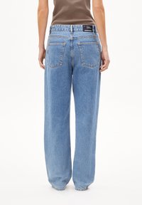 Person som bär hög midja, ljusblå raka jeans med bakfickor och en brun ärmlös topp, stående mot en vit bakgrund.