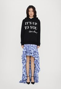 Svart hoodie med vit text "DET ÄR UPP TILL DIG" som bärs över en blå rutmönstrad volangkjol. Svarta klackskor fulländar outfiten.