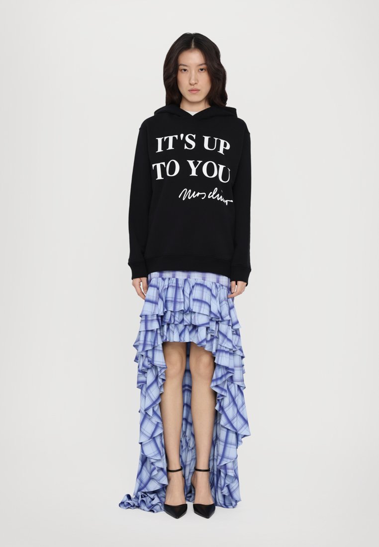 Un sweat à capuche noir avec le texte blanc "C'EST À VOUS DE DÉCIDER" porté par-dessus une jupe plissée à carreaux bleus. Des chaussures à talons hauts noires complètent la tenue.