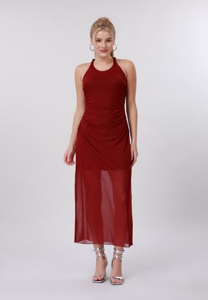 Nő, aki ujjatlan bordó, nyakba akasztós ruhát visel átlátszó szoknyával és ezüst pántos magas sarkú szandálban, egyszerű háttér előtt áll.