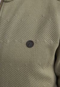 La tela texturizada verde oliva presenta un nervio diagonal y una insignia redonda negra en la zona del pecho. El diseño es moderno y funcional.