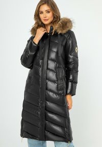 Schwarzer, gepolsterter langer Mantel mit glänzender Oberfläche. Verfügt über eine mit Fell gefütterte Kapuze, Reißverschlusstaschen und strukturierte Paneelakzente. Silberne Hardware-Details.