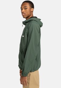 Leichte grüne Jacke mit Kapuze, elastischen Bündchen und einer Fronttasche. Sie verfügt über ein kleines Logo auf der Brust und eine glatte Textur.