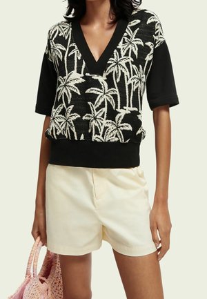 Nainen pukeutunut mustavalkoiseen palmuaiheiseen V-mekkoon, jossa mustat hihat, beigehousuihin, pitäen vaaleanpunaista punottua laukkua.