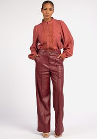 Rostfarbene Bluse mit gestickten Details und Rüschenkragen, kombiniert mit hoch taillierten bordeauxfarbenen Lederhosen mit weitem Bein.