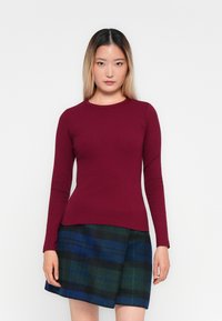 Pitkähihainen burgundinpunainen ribbipaita, jossa on pyöreä kaula-aukko, yhdistettynä ruudulliseen hameeseen, jossa on vihreitä, sinisiä ja mustia sävyjä. Sujuva pinta.