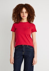 Νεαρή γυναίκα με σγουρά μαλλιά, που φοράει κόκκινο κοντομάνικο πουκάμισο και σκούρο μπλε τζιν, στέκεται μπροστά σε απλό φωτεινό φόντο.