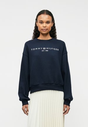 Žena s dugom kovrčavom kosom u tamnoplavom Tommy Hilfiger džemperu i bijeloj plisiranoj suknji stoji pred jednobojnom pozadinom.