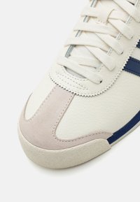 Hvide lædersneakers med en lys grå ruskindsdetalje. Har blå striber, struktureret gummisål og flade bomulds snørebånd.