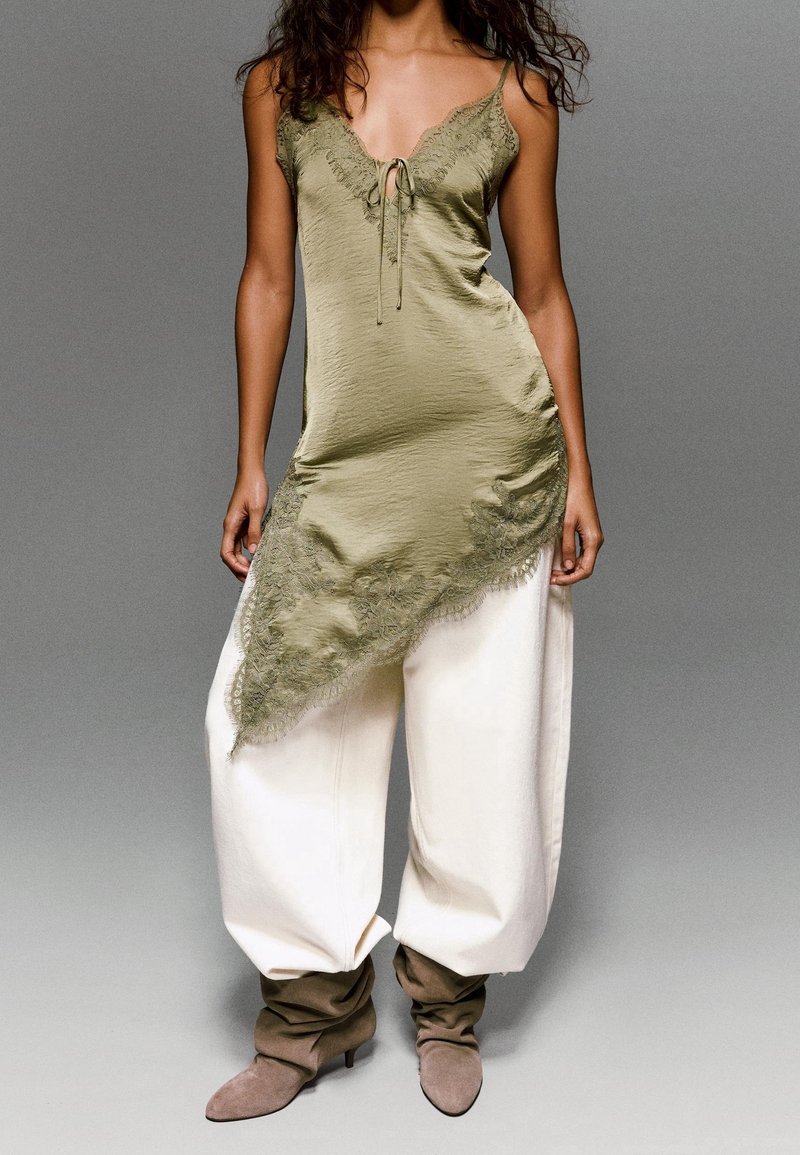 Femme portant un caraco asymétrique vert olive orné de dentelle, un pantalon blanc ample et des bottes en daim taupe décontractées sur un fond gris uni.
