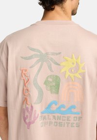 T-shirt rose clair avec divers graphismes colorés et texturés comprenant un palmier, un soleil, un visage, des vagues, du corail et le texte "ÉQUILIBRE DES OPPOSÉS."