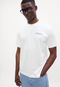Tricou alb din bumbac cu mâneci scurte, guler rotund și logo brodat negru. Purtați cu șorturi din denim bleu deschis. Ochelari de soare mari, negri.
