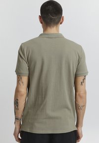 Olivengrønn polo-skjorte laget av teksturert stoff, med krage og korte ermer. Synlige tatoveringer på underarmene.