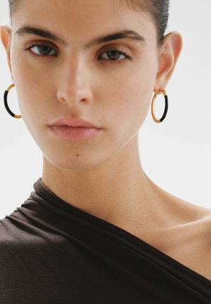 Pendientes de aro en oro y negro adornan a una modelo, con un acabado metálico suave y una forma redonda. La modelo lleva una blusa oscura con caída.