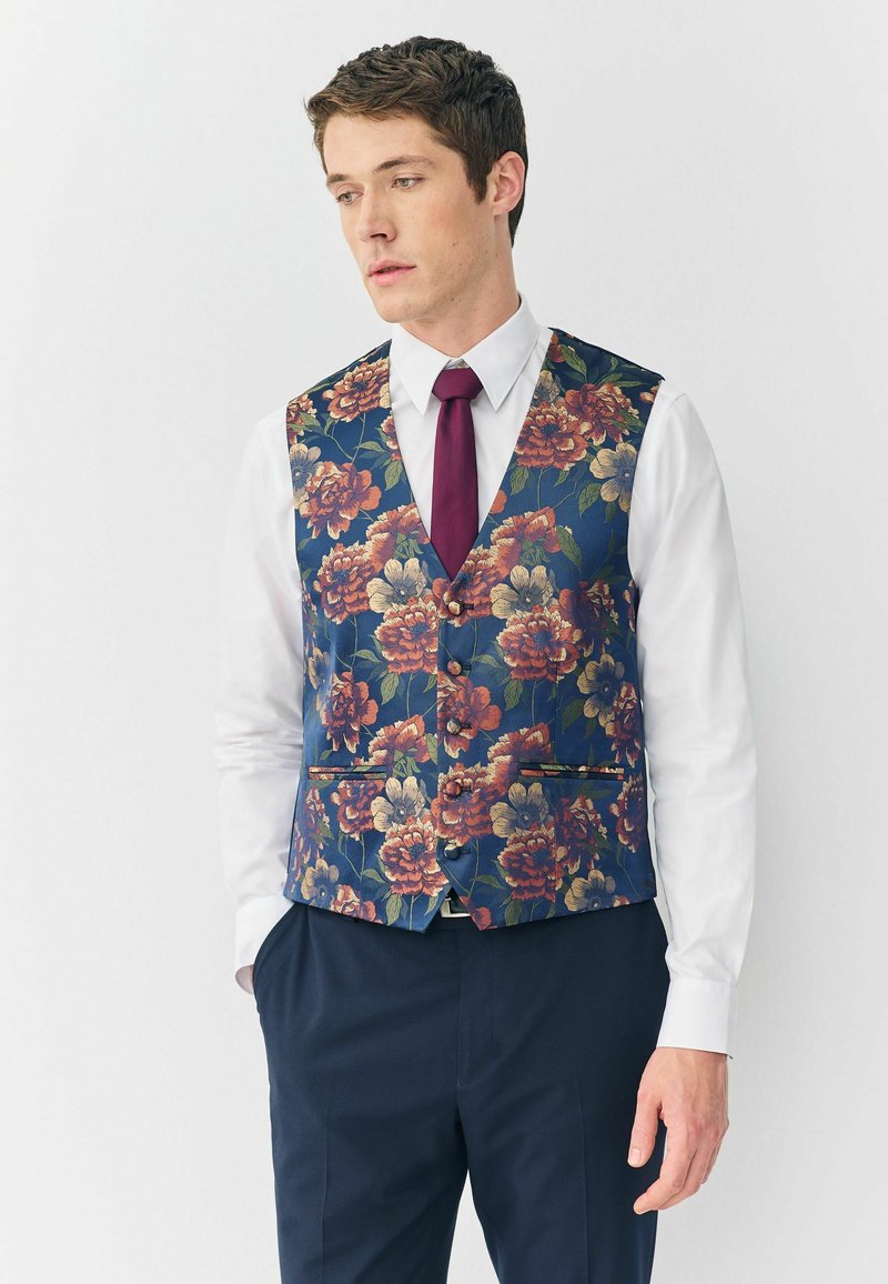 Navy Weste mit floralem Muster und großen orange- und gelbblühenden Motiven. Mit spitzem Kragen, Knöpfen und Seitentaschen; über einem weißen Hemd gestylt.