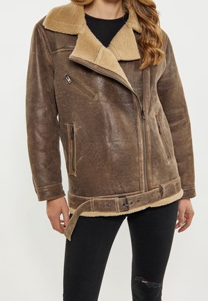 Frau trägt braune Lammfelljacke mit beigem Fleece-Kragen, Reißverschlusstaschen und tailliertem Gürtel, kombiniert mit schwarzen, zerrissenen Skinny-Jeans.