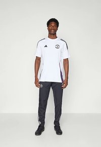 Witte sportieve t-shirt met zwarte en paarse accenten, voorzien van een geborduurd logo en drie strepen op de schouders, gecombineerd met een donkere broek.