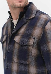 Marinblå och brun rutig skjortjacka i en strukturerad ullblandning, med dragkedjestängning, två bröstfickor och krage. Knapparna är mörka och runda.