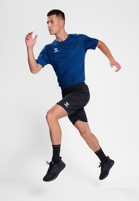 Kék sportpóló és fekete rövidnadrág, elegáns szabással. Fekete cipők, texturált talppal. A modell futó pózban áll egy egyszerű háttér előtt.