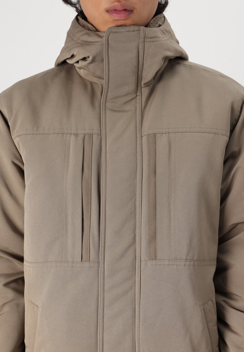 Beżowa ocieplana kurtka z wysokim kołnierzem, poziomymi szwami i dwiema pionowymi kieszeniami na zamek na piersi. Miękka faktura i elegancki design.