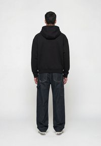 Άντρας που στέκεται με την πλάτη προς τα εμπρός, φοράει μαύρο φούτερ με κουκούλα, σκούρο, άνετο τζιν με πλαϊνές τσέπες και λευκά αθλητικά παπούτσια, σε απλό ανοιχτό φόντο.