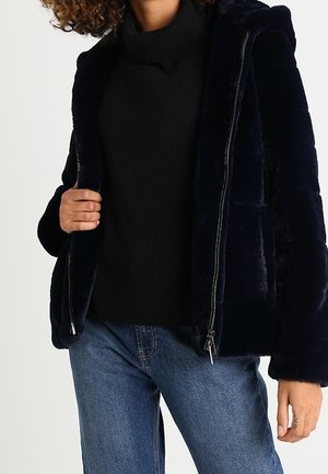 Persoon die een zwarte coltrui, een donkerblauwe nepbontjas met ritssluiting en een blauwe spijkerbroek draagt, staand tegen een witte achtergrond.