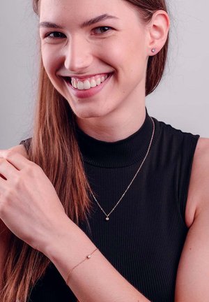 Rose gold necklace and bracelet with small spherical accents, worn on a model with a black ribbed turtleneck, smiling against a neutral background.