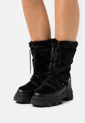 Botas negras hasta la mitad de la pantorrilla con suelas gruesas y textura de piel, llevadas por una persona con las piernas al aire sobre un fondo blanco.
