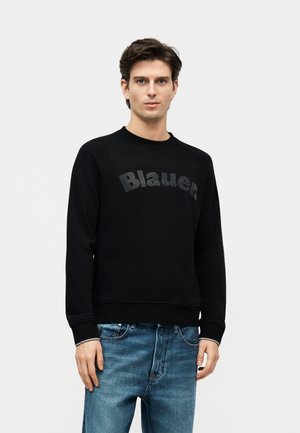 Fiatal férfi fekete "Blauer" feliratú pulóverben és kék farmert visel, egyszerű fehér háttér előtt áll.