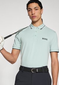Svetlozelené polokošeľa s čiernymi detailmi, s logom „BOSS“. V kombinácii s tmavomodrými nohavicami a čiernym tkaným opaskom. Drží golfovú palicu.