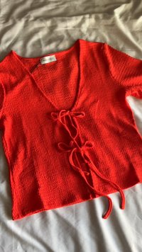 Topo curto vermelho de mangas curtas com decote em V e detalhe de atar na frente. Tecido texturizado com um corte solto, apresentando laços finos e ajustáveis.