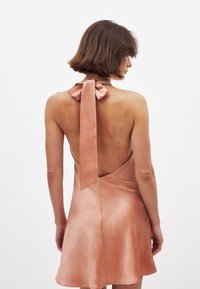 Satin-Halterkleid in sanftem Pfirsich mit offenem Rücken. Es verfügt über eine gebundene Schleife am Hals und einen fließenden Rock mit glatter Textur.