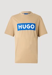 Béžové bavlnené tričko s modrým obdĺžnikovým logom s nápisom „HUGO" v bielych písmenách, okrúhlym výstrihom a krátkymi rukávmi. Hladká textúra.