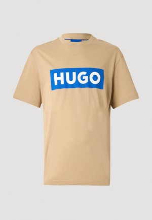 Beige cotton T-shirt featuring a blue rectangular logo with "HUGO" in white letters, crew neck, and short sleeves. Smooth texture.
