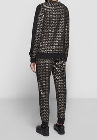 Svart träningsoverall med ett mönster av upprepande guld EA7-logotyp. Överdel med långa ärmar och manchetter; byxorna har elastiska muddar.