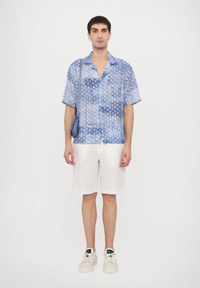 Chemise à manches courtes à motifs bleus, shorts blancs, baskets blanches. Tissu léger avec des boutons. Silhouette décontractée, sac minimaliste en bandoulière.
