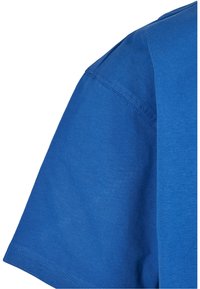 Tricou din bumbac albastru, cu mâneci scurte, textură netedă și detalii de cusătură vizibile pe umeri și pe zona mânecilor.