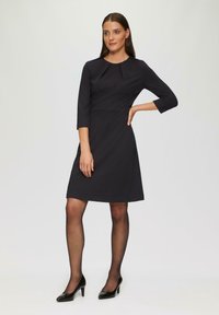 Robe noire à manches trois-quarts, avec un détail plissé au niveau du col, un corsage ajusté et une jupe évasée. Portée avec des talons noirs.