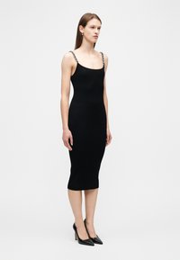 Γυναίκα που φοράει εφαρμοστό μαύρο μεσομήκι φόρεμα με λεπτούς ιμάντες αλυσίδας και μαύρα ψηλοτάκουνα παπούτσια με μυτερή μύτη, στέκεται μπροστά σε απλό λευκό φόντο.