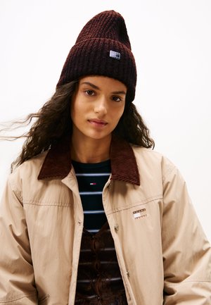 Beanie a coste color bordeaux con punta e patch con logo, indossato sopra una giacca beige con colletto in cotone e una maglietta a righe sotto.