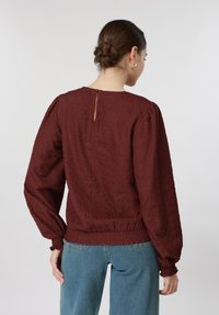 Γυναίκα με καστανά μαλλιά μαζεμένα σε κότσο, που φοράει χρυσούς κρίκους στα αυτιά, μια μπλούζα μακρυμάνικη μπορντό με υφή και μπλε τζιν, φαίνεται από πίσω.