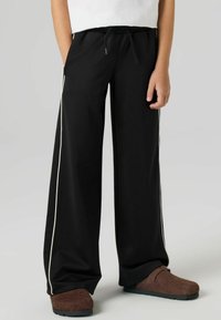 Svarte joggebukser med vide ben og snor i midjen, med hvite side-striper. Laget av mykt stoff, designet for komfort og enkelhet.