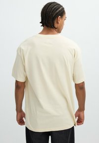 Persona con el pelo trenzado que lleva una camiseta beige holgada de manga corta y pantalones negros, de pie de espaldas frente a un fondo liso.